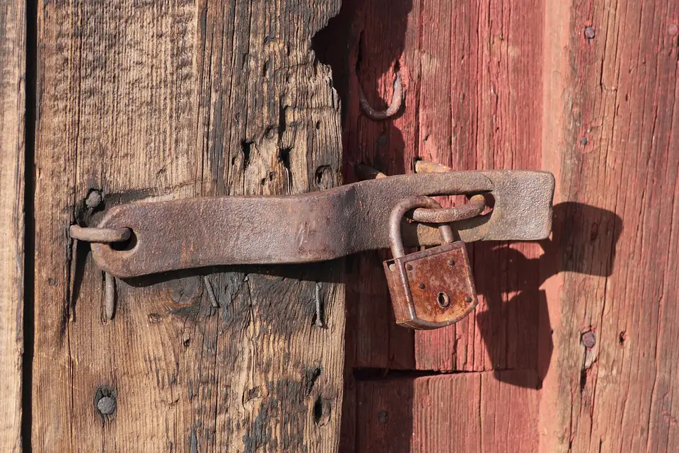A small padlock securing a rusty iron hasp on a weathered red wooden door