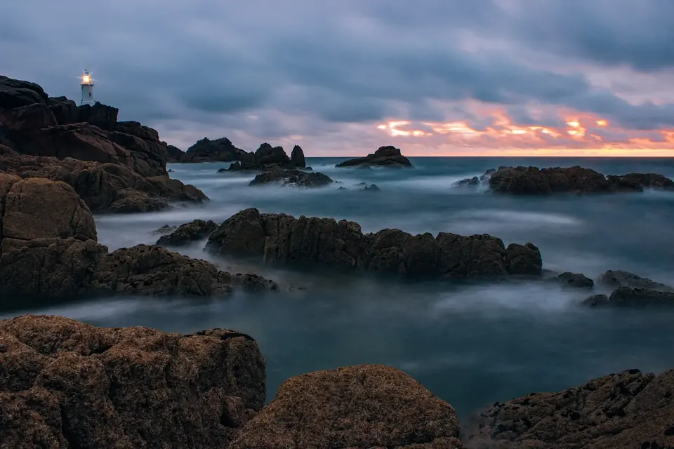 A small white lighthouse lit at dusk on a rocky headland, above a misty sea and dark rocks under a cloudy sky